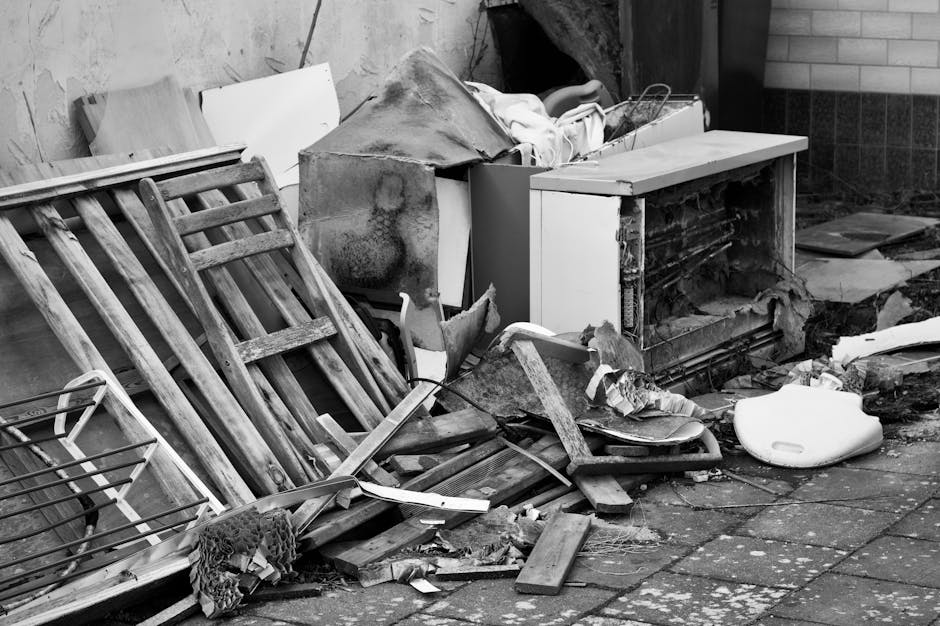 A black and white photograph depicting a cluttered outdoor scene with various discarded items piled against a wall. In the foreground, several wooden slatted garden chairs are broken and stacked hapha