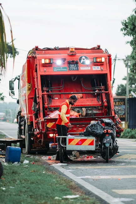 A worker wearing a red and yellow high-visibility vest is engaged in rubbish collection beside a large red waste management truck parked on the side of a street. The truck's rear opening is raised, re