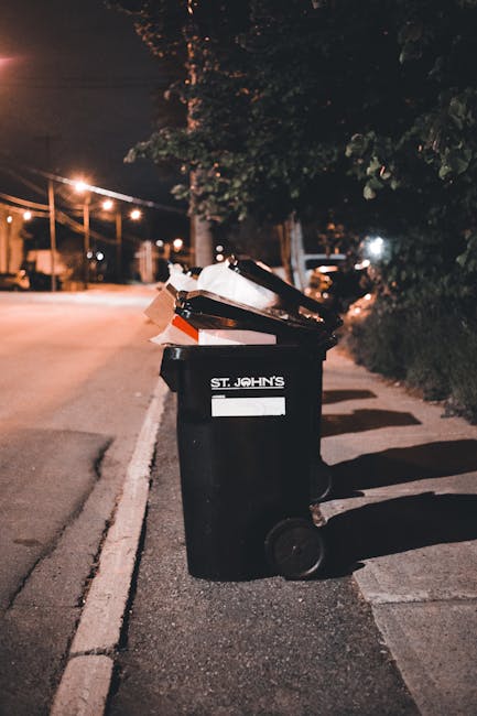The image depicts several large trash bags, made of black plastic and some with visible text and branding, placed along the curbside of a paved street. The bags appear to contain discarded household o